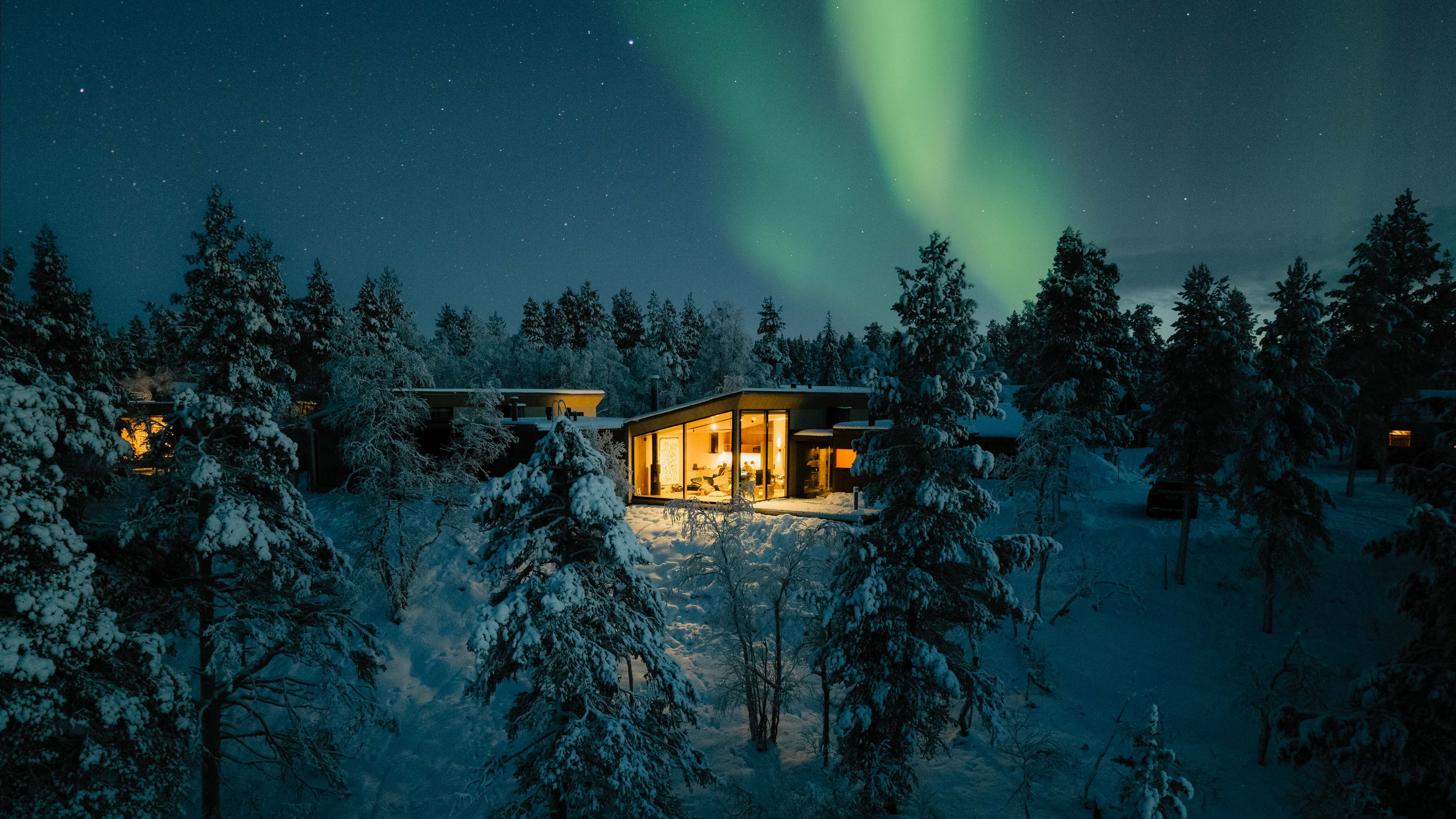 A small lit-up cabin in a snowy woodland under the Aurora Borealis